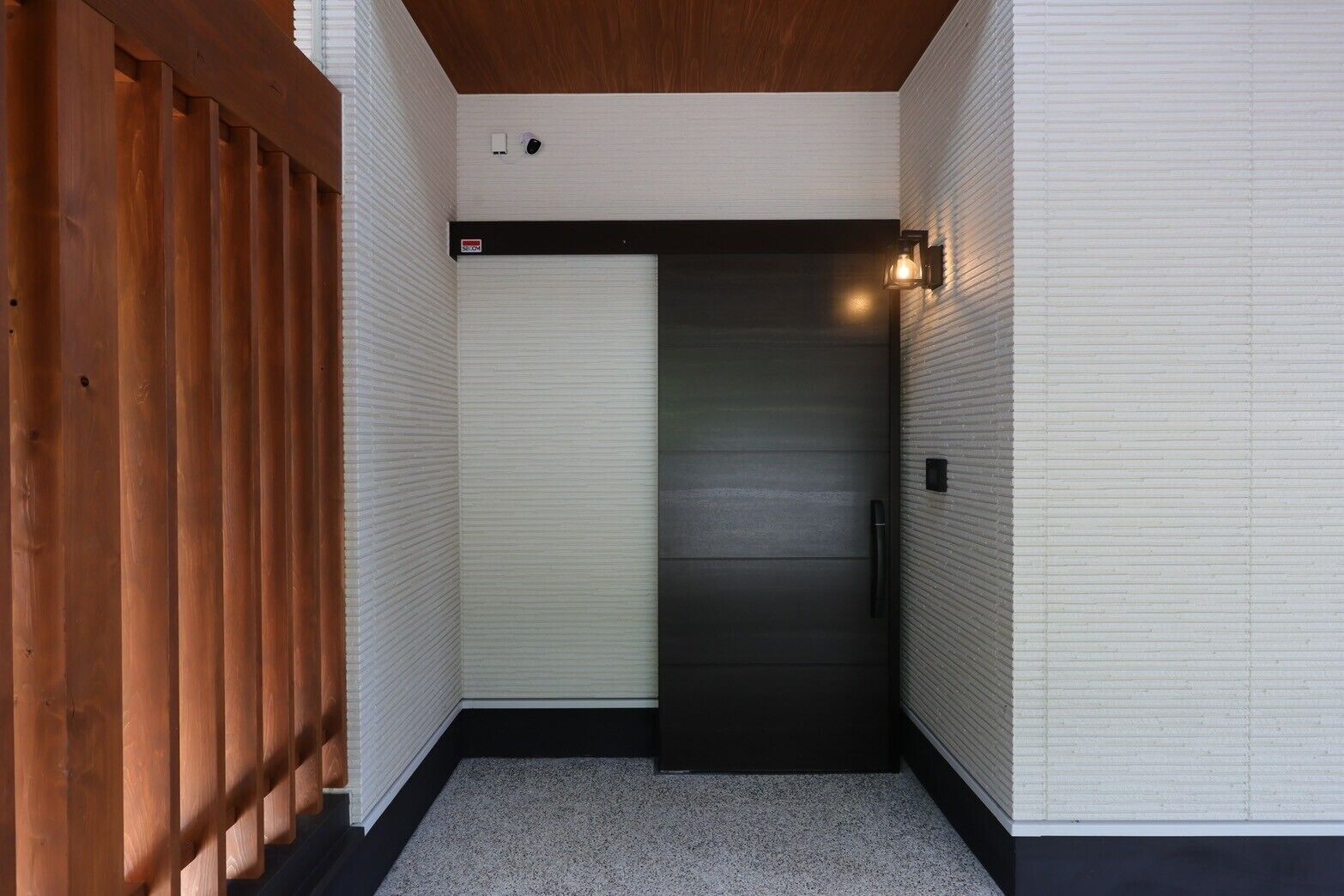 A narrow hallway with a wooden fence on one side, a black door with a handle, and a light fixture on the wall.