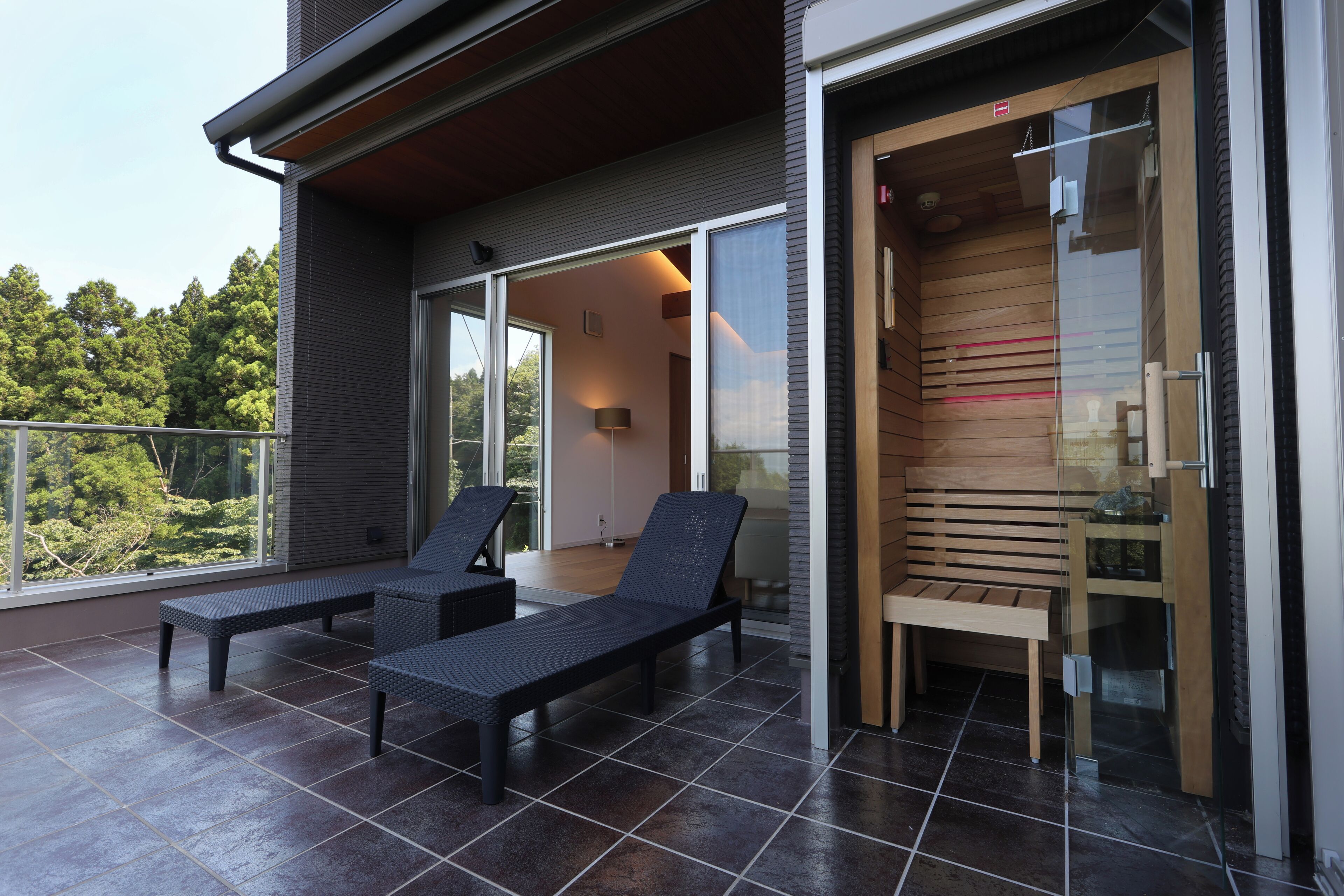 A balcony with two lounge chairs and a small table, a sliding glass door leading to a wooden interior, and a view of trees.