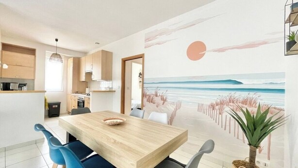 A modern kitchen with wooden cabinets, a dining area with a wooden table and blue chairs, and a mural of a beach scene on the wall. - Le Benodet Centre. In the center of town 200m from the beach (Bénodet)