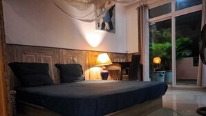Een slaapkamer met een bed, een nachtkastje met een lamp, een stoel en een groot raam met uitzicht op groen.