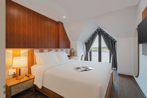 Habitación de hotel con una cama grande, una mesita de noche con lámpara y otra mesita con teléfono, además de una ventana con cortinas.