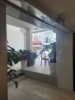 Pasillo con puerta de cristal que da a un área exterior con plantas en macetas y una silla. Se observa un letrero que indica una salida.