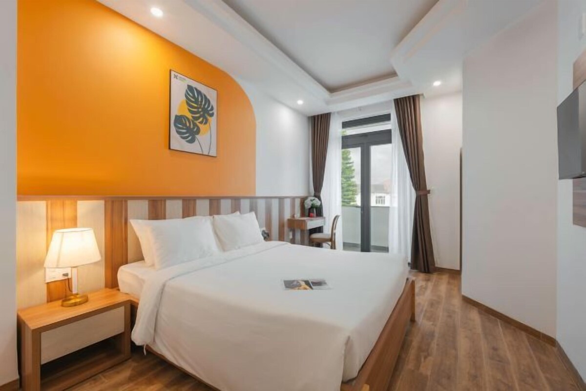 A hotel room with a bed, a nightstand with a lamp, a painting on the wall, and a view of the outdoors through a window.