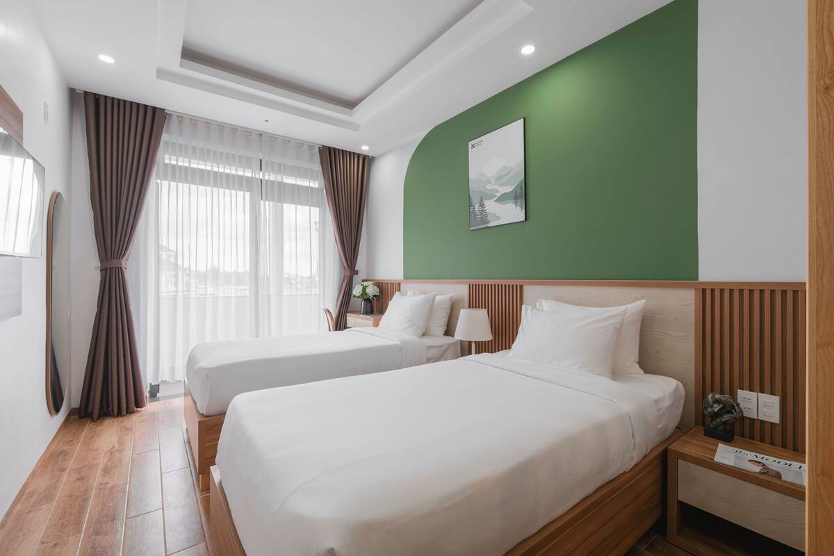 Habitación de hotel con dos camas, una pared de acento verde, un cabecero de madera y una mesita de noche con una lámpara y una revista.
