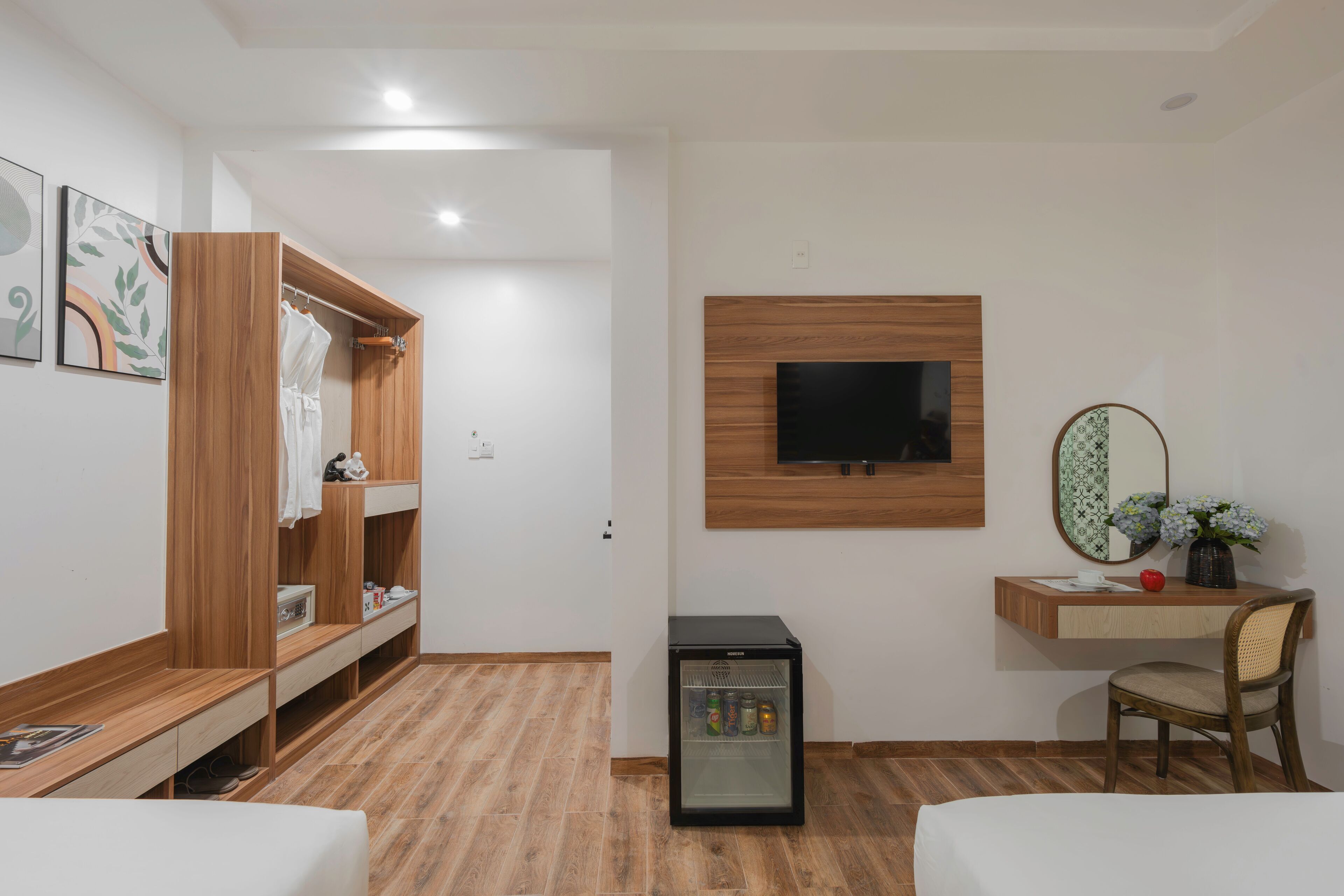 A hotel room with a wooden wardrobe, a small fridge, a desk with a chair, a TV mounted on the wall, and a mirror.