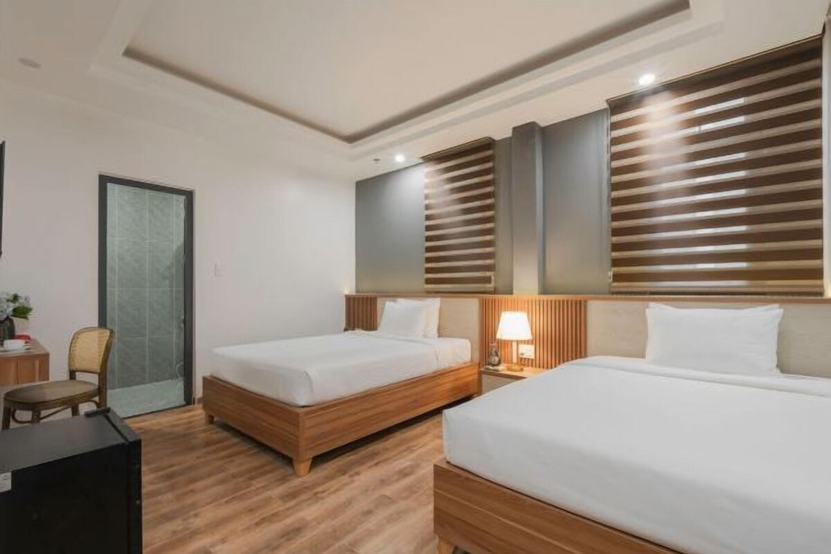 A hotel room with two single beds, a wooden floor, a desk with a chair, and a large window with blinds.
