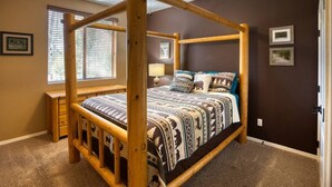 A four-poster bed with a patterned bedspread, a bedside table, a lamp, and a window with blinds.