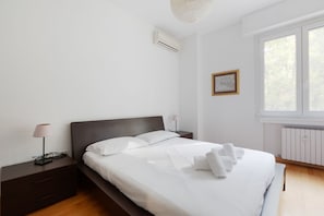 Un dormitorio moderno con cama, mesitas de noche, lámpara, equipo de aire acondicionado, un cuadro enmarcado y una ventana.