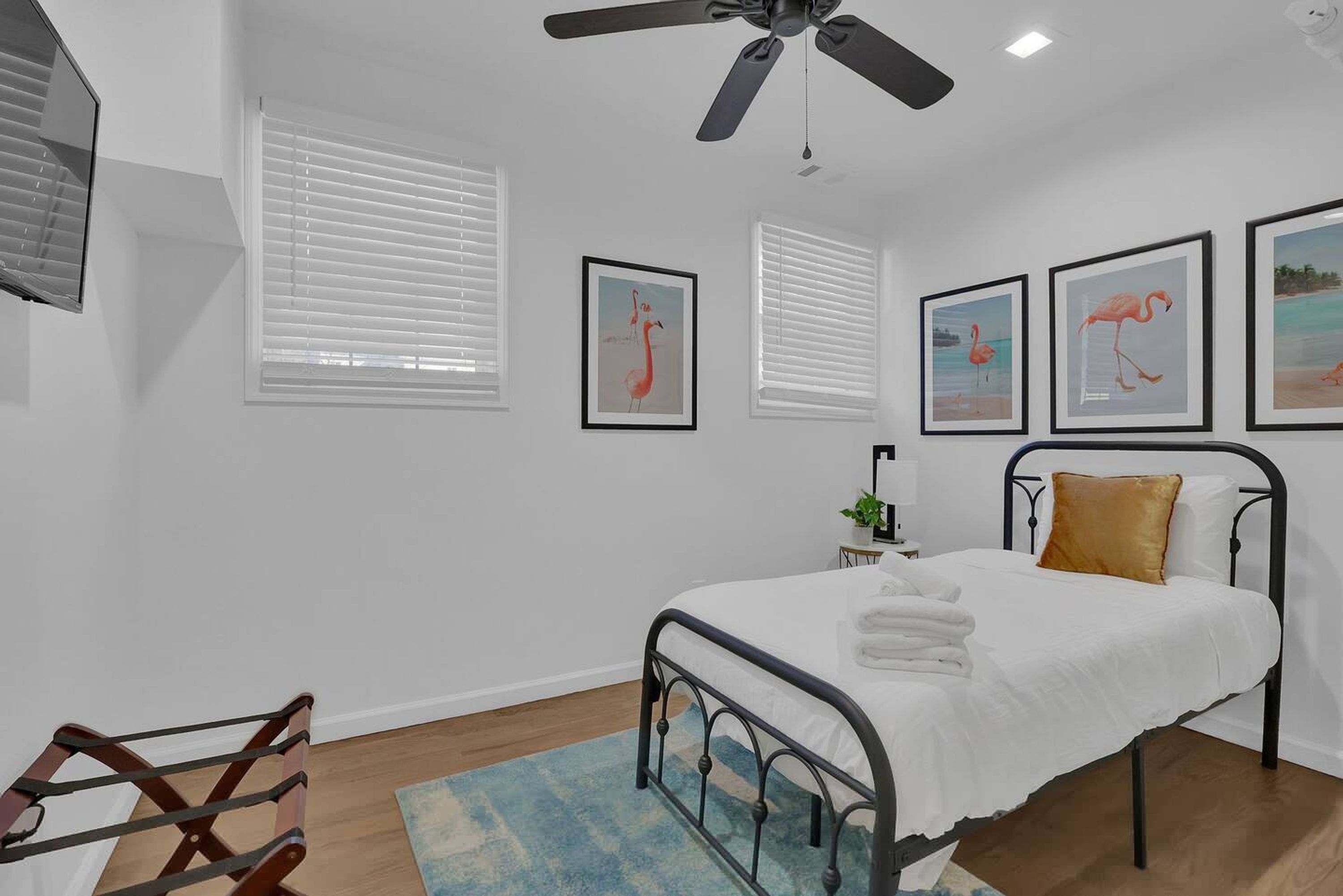 Quarto com cama, ventoinha no teto, paredes brancas e quadros com flamingos.