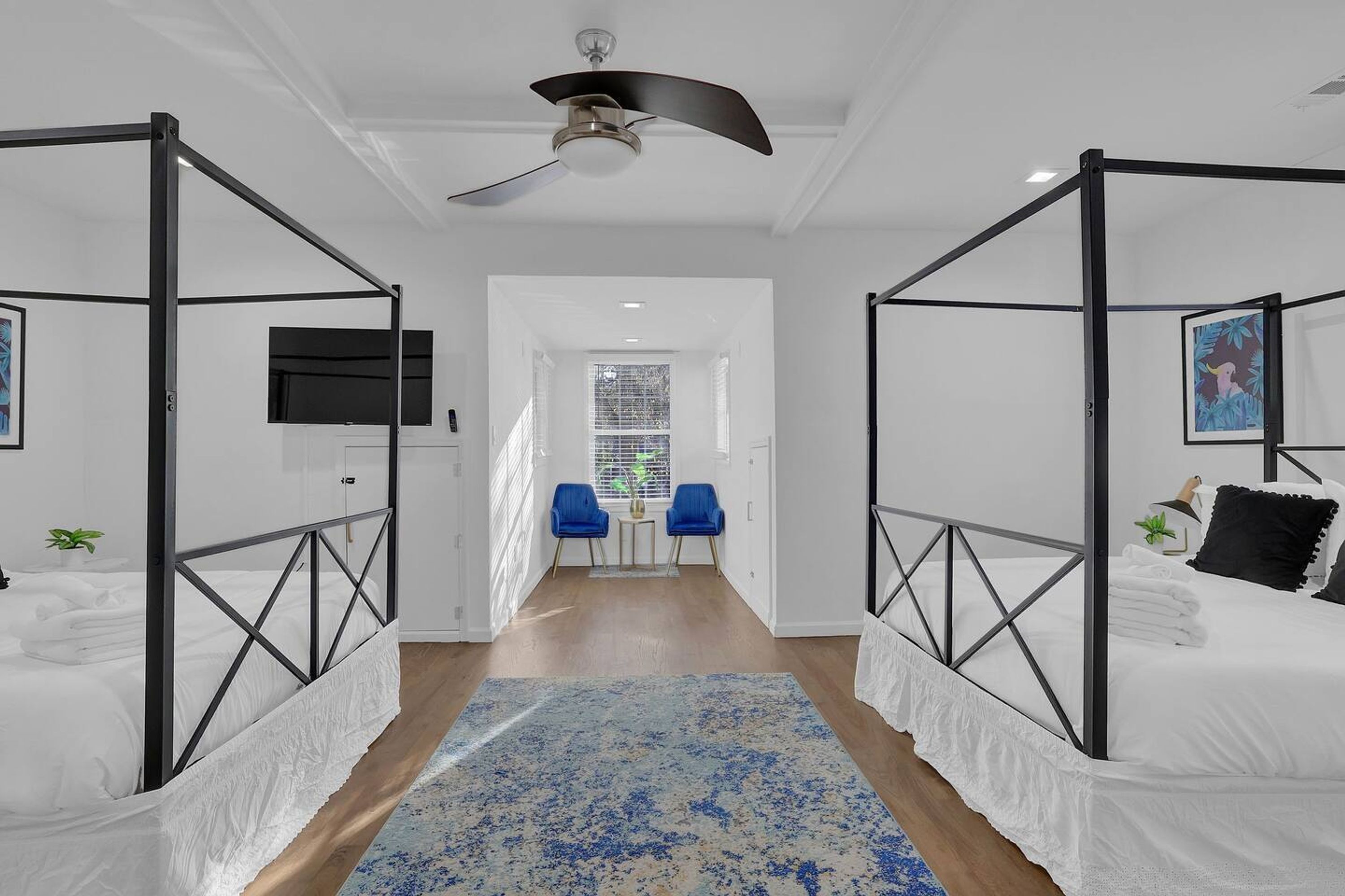 A modern bedroom with a four-poster bed, a ceiling fan, and a view to a hallway with two chairs and a window.