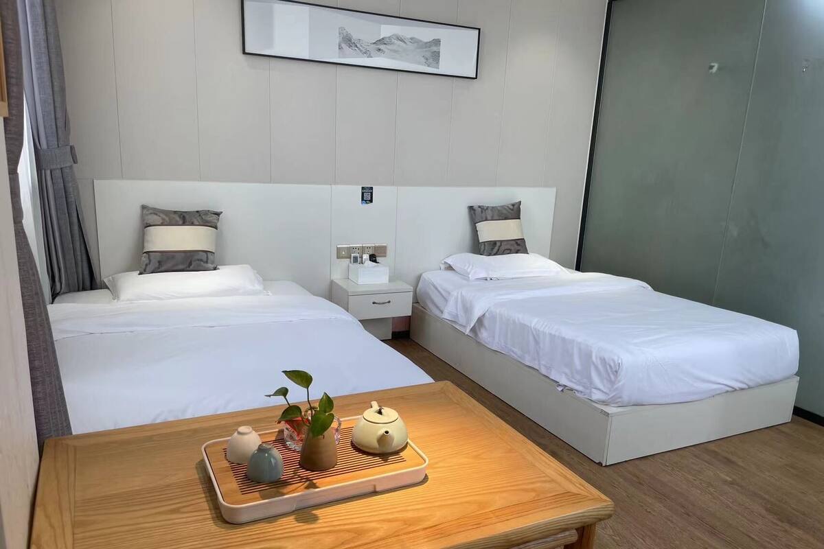A hotel room with two beds, a bedside table with a tray of tea and a plant, and a framed picture on the wall.