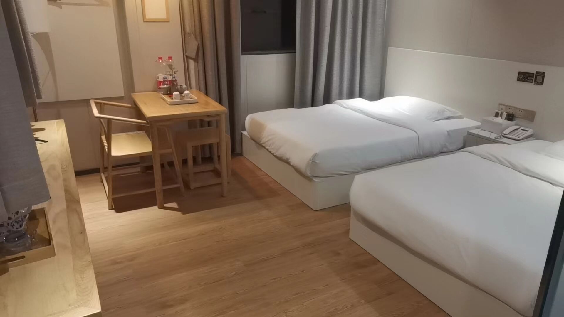 A hotel room with a wooden desk, two beds, a small table with a tray, and a window with curtains.