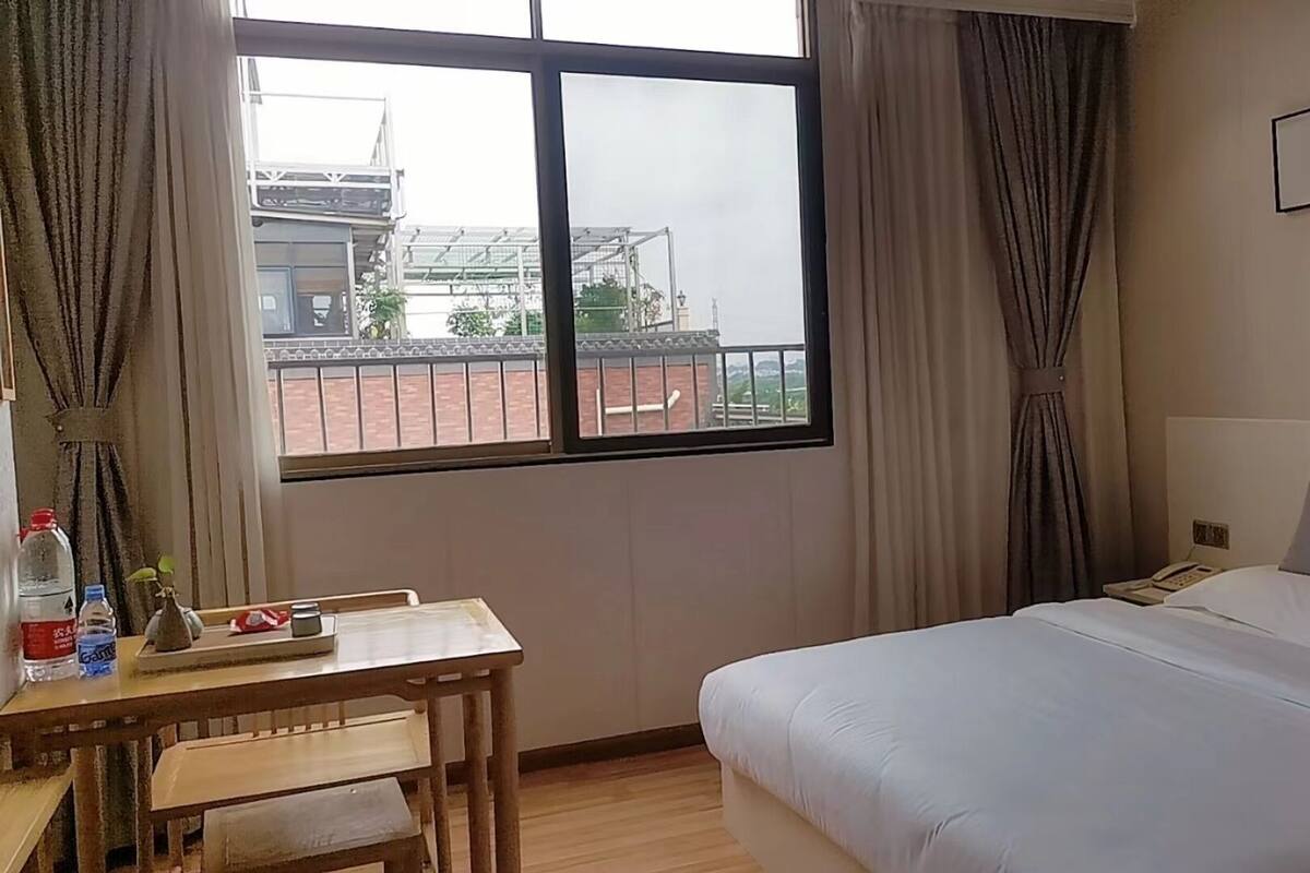 A hotel room with a bed, a desk with a chair, a window with curtains, and a view of a building outside.