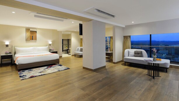 A modern hotel room with a large bed, a sofa, a dining area, and a view of the city at dusk. - Ginger Hyderabad Airport Road (Hyderabad)