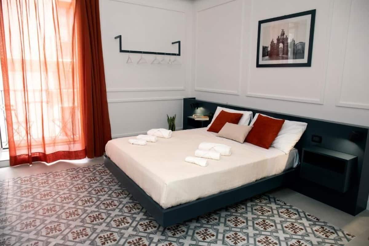 A neatly made bed with towels, a bedside table, a wall-mounted shelf, and a framed picture.
