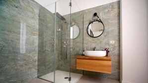Un baño moderno con ducha de vidrio, pared de mármol, tocador de madera y espejo redondo.