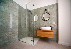 Un bagno moderno con doccia a vetro, parete in marmo, mobile contenitore in legno e specchio tondo.