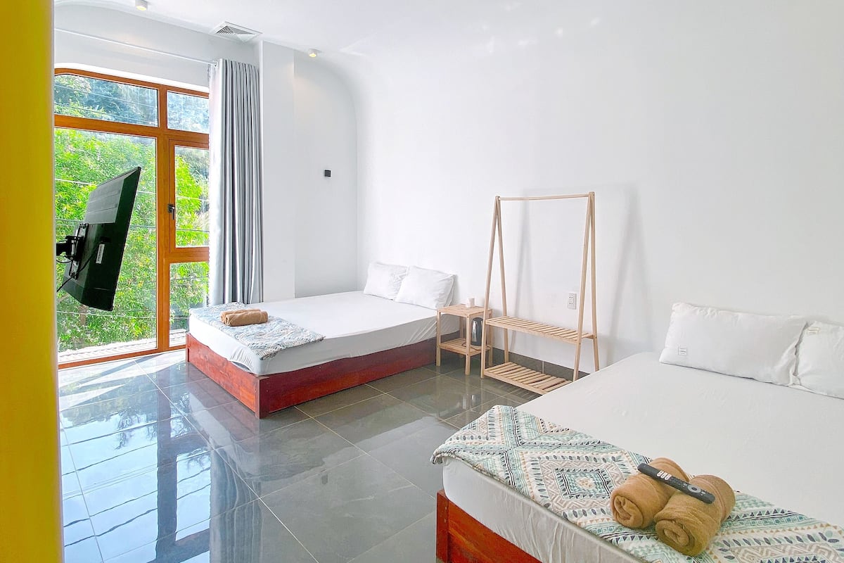 A hotel room with two beds, a wooden shelf, and a view of greenery through the window.