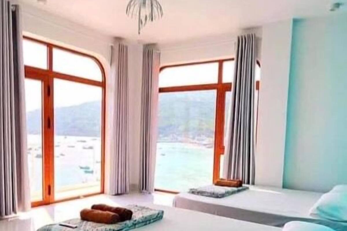 A modern bedroom with a large bed, two windows with curtains, and a view of the sea.