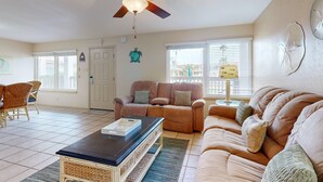 A living room with a ceiling fan, a brown leather sofa, a white couch, and a dining area with wicker chairs. - Beach Access Across the Street, Shared Heated Pool, Pet Friendly (Corpus Christi)