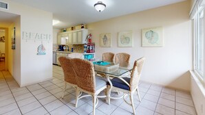 A dining area with wicker chairs and a glass table, a kitchen in the background, and a wall with a 'BEACH' sign. - Beach Access Across the Street, Shared Heated Pool, Pet Friendly (Corpus Christi)