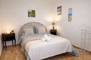 Cama bem arrumada com edredom branco e duas almofadas, mesa de cabeceira com abajur, e um varal fixado na parede.