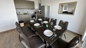 A modern dining area with a dark wooden table set for six, surrounded by leather chairs. A kitchen with grey cabinets and a refrigerator is visible in the background.