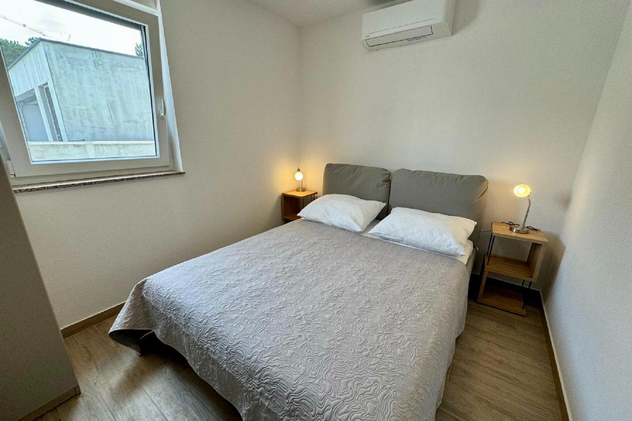A bedroom with a bed, two bedside tables, a window, and an air conditioning unit.