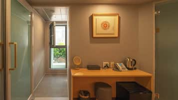 A compact hotel room with a wooden desk, a framed artwork, a kettle, a telephone, and a small stool.