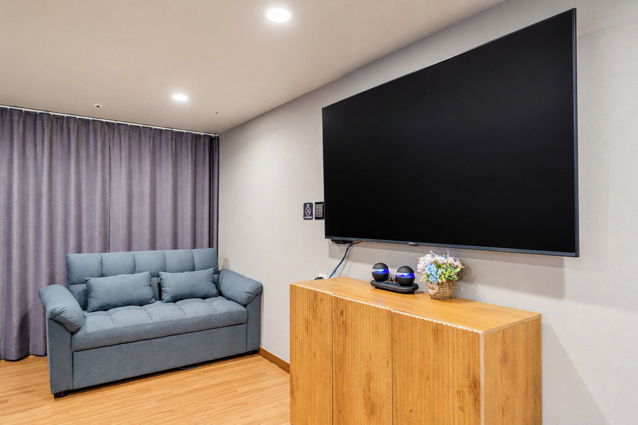 A modern living room with a grey sofa, a wooden cabinet, and a large flat-screen TV mounted on the wall.