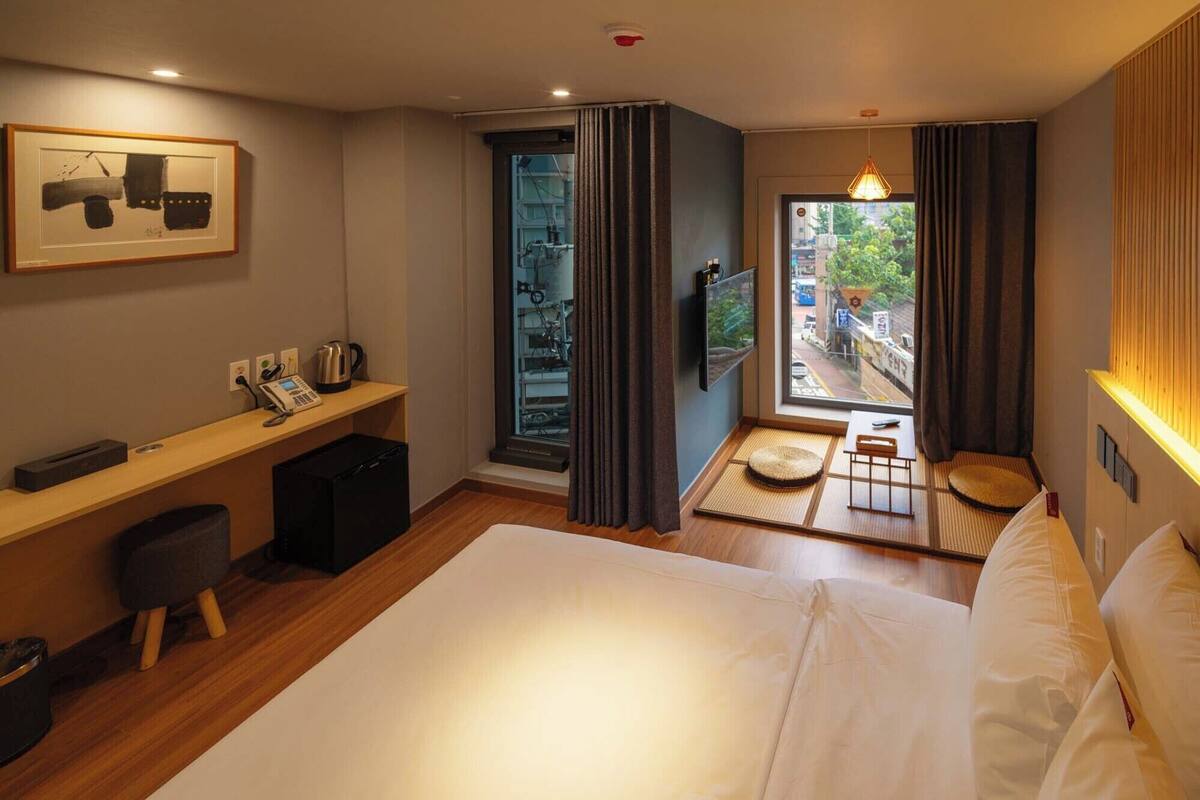 Une chambre d’hôtel avec un grand lit, un bureau avec un téléphone et une bouilloire, une télévision et une vue sur la rue par la fenêtre.