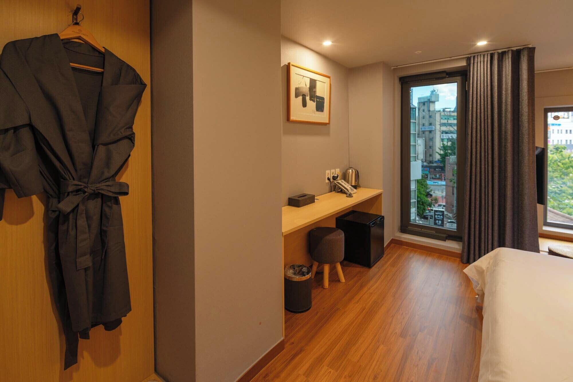 Habitación de hotel con piso de madera, una cama, un escritorio con silla, un cesto de basura, una bata colgada en una puerta de madera y una ventana con cortinas.