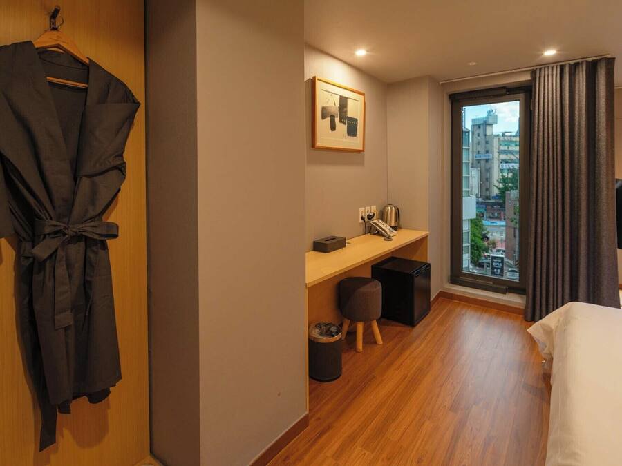 Habitación de hotel con piso de madera, una cama, un escritorio con silla, un cesto de basura, una bata colgada en una puerta de madera y una ventana con cortinas.