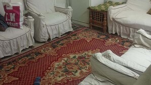 A room with two white-covered chairs, a small wooden table, a refrigerator, and a patterned rug. - Beautiful Apartment with 2 Bdr and 2 Living Rooms (Ard El lewa, El Mohandessin)