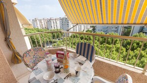 A balcony with a table set for two, featuring a patterned tablecloth, a bottle of orange juice, and a box of B-SAN. The balcony overlooks a residential area with greenery and buildings in the background. - Apartment Nube in Grau i Platja (Gandia)