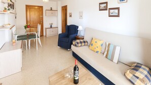 A living room with a sofa, a dining table with chairs, a wooden coffee table with a bottle of wine, and a blue armchair. - Apartment Nube in Grau i Platja (Gandia)