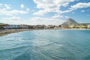 A coastal beach scene with a mountain in the background, buildings along the shore, and people on the beach. - Apartment Zeus in Platja de l Arenal (Javea)