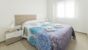 A neatly made bed with a patterned bedspread, a bedside table with a lamp, and a window with blinds. - Apartment Egeu in D nia (Denia)