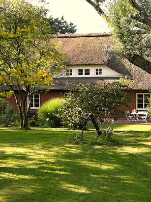 A house with a thatched roof, surrounded by a well-maintained garden with a lawn and a small table and chairs.