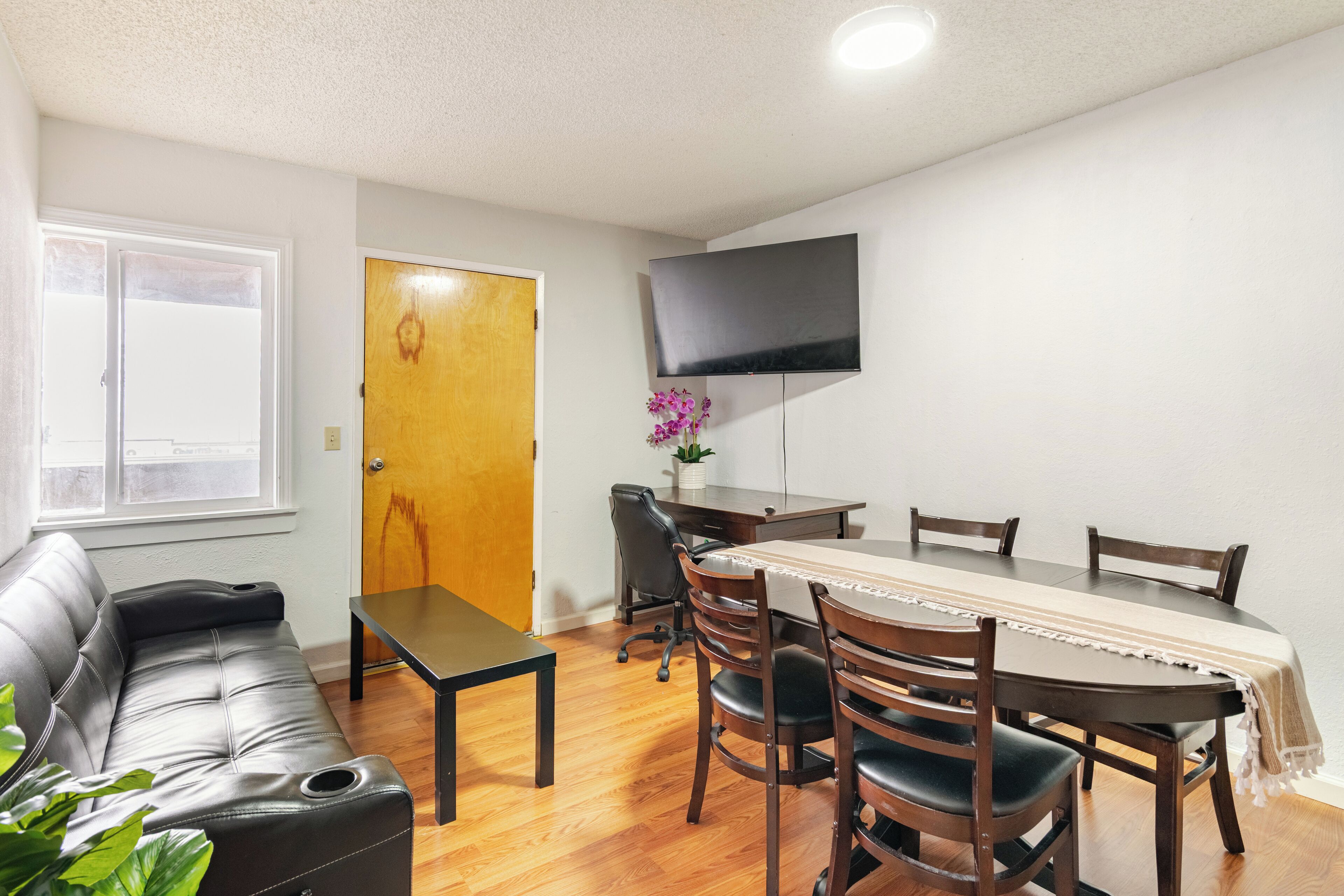 A living room with a sofa, a dining table with chairs, a TV mounted on the wall, and a door.