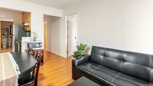 Una sala de estar moderna con un sofá de cuero negro, una mesa de comedor de madera y sillas. Se ve una cocina al fondo con un microondas y armarios.