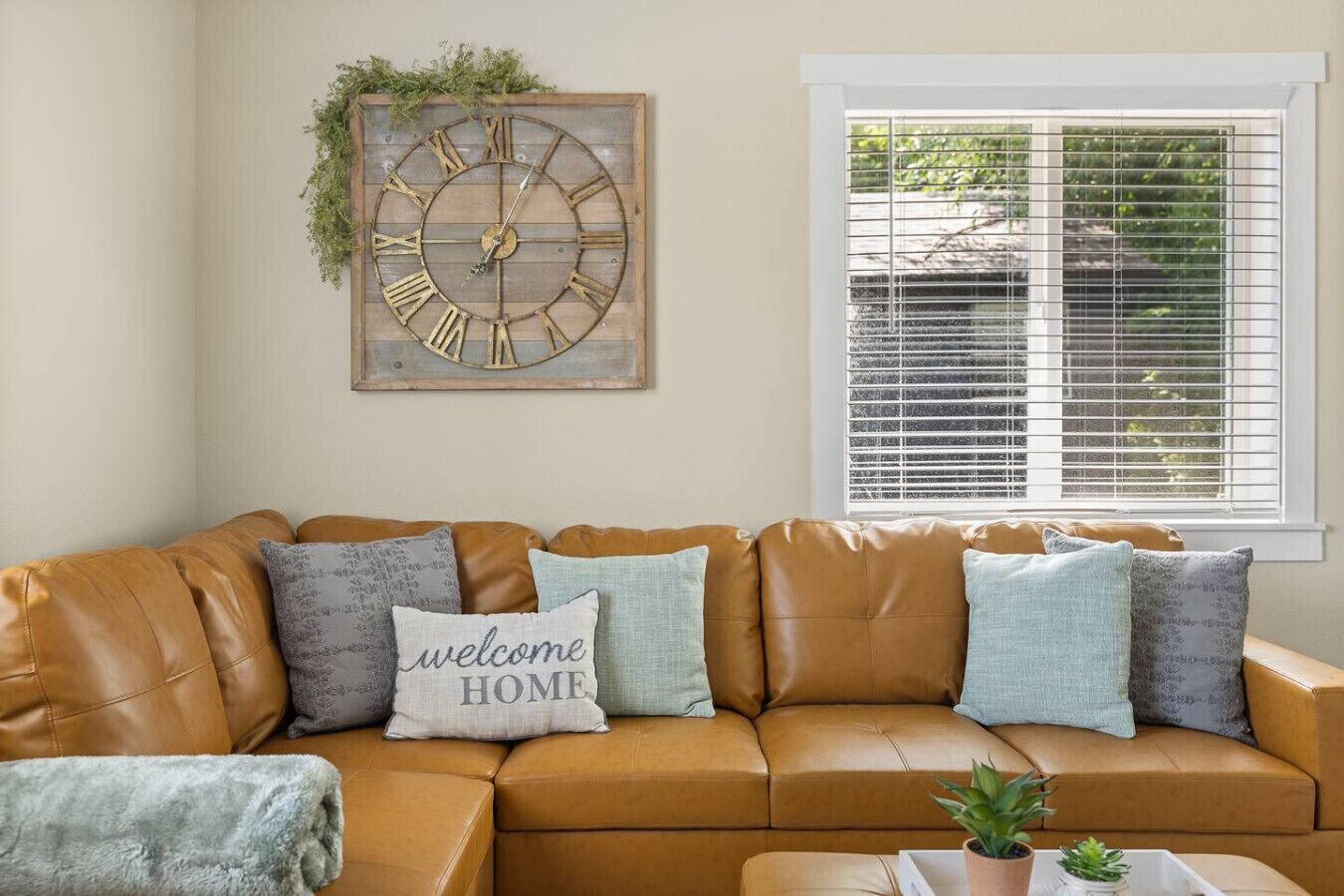 Ein beiges Ledersofa mit 'willkommen ZUHAUSE'-Kissen, eine rustikale Wanduhr und ein Fenster mit Jalousien.