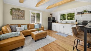 A modern living room with a sofa, ottoman, and a coffee table. There is a kitchen area with white cabinets, a refrigerator, and a stove. - Crow peak Cabin: Peaceful stay (Spearfish)