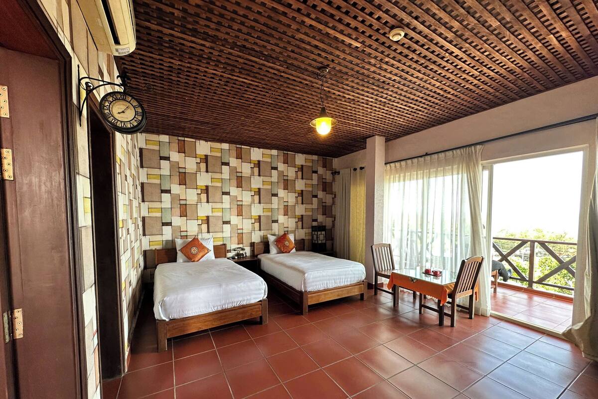 A room with two beds, a wooden ceiling, a wall clock, a table with chairs, and a balcony with a view.