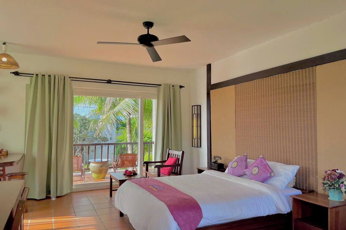 A hotel room with a bed, a chair, a table, and a balcony with a view of greenery.