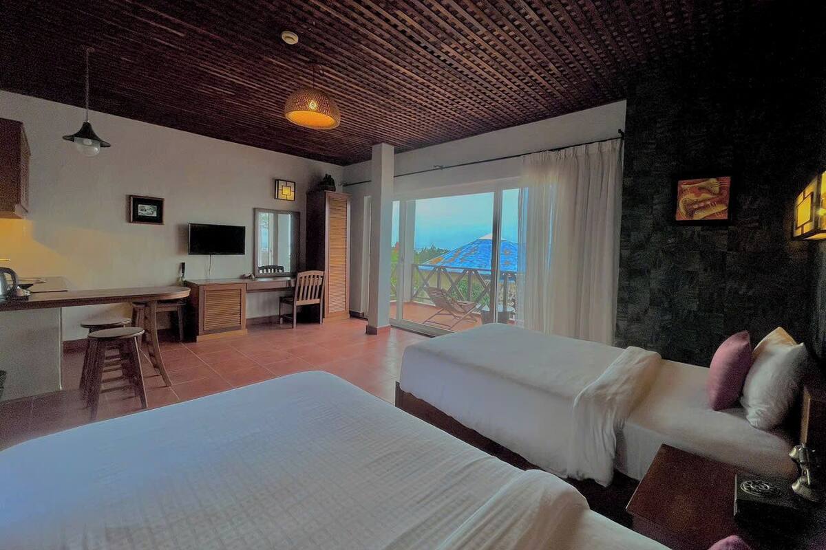 A hotel room with two beds, a desk with a chair, a TV, and a balcony with a view.