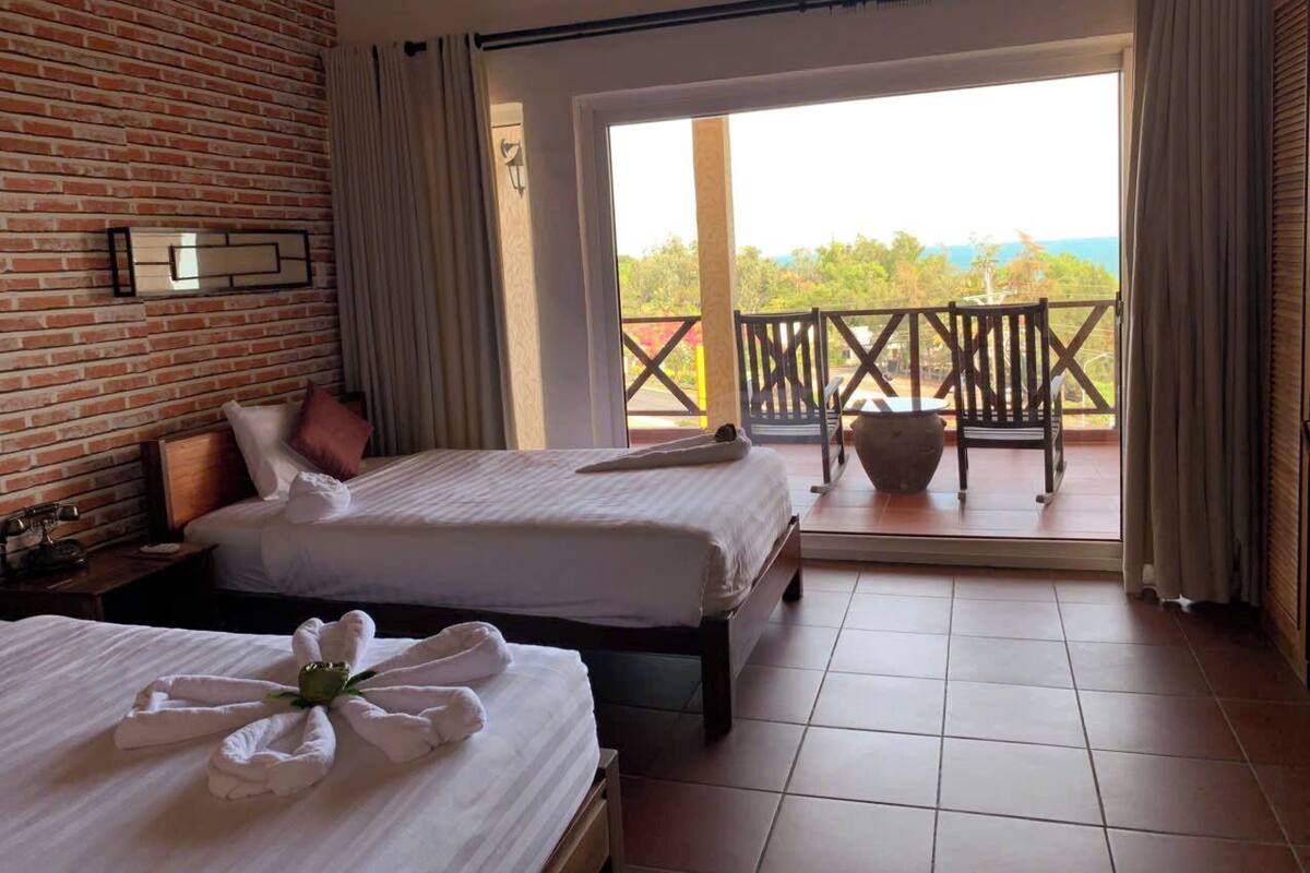 A hotel room with two beds, a brick wall, a balcony with a view, and a large window.