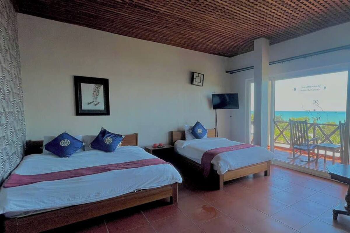 A hotel room with two beds, a TV, and a balcony with a view of the sea.