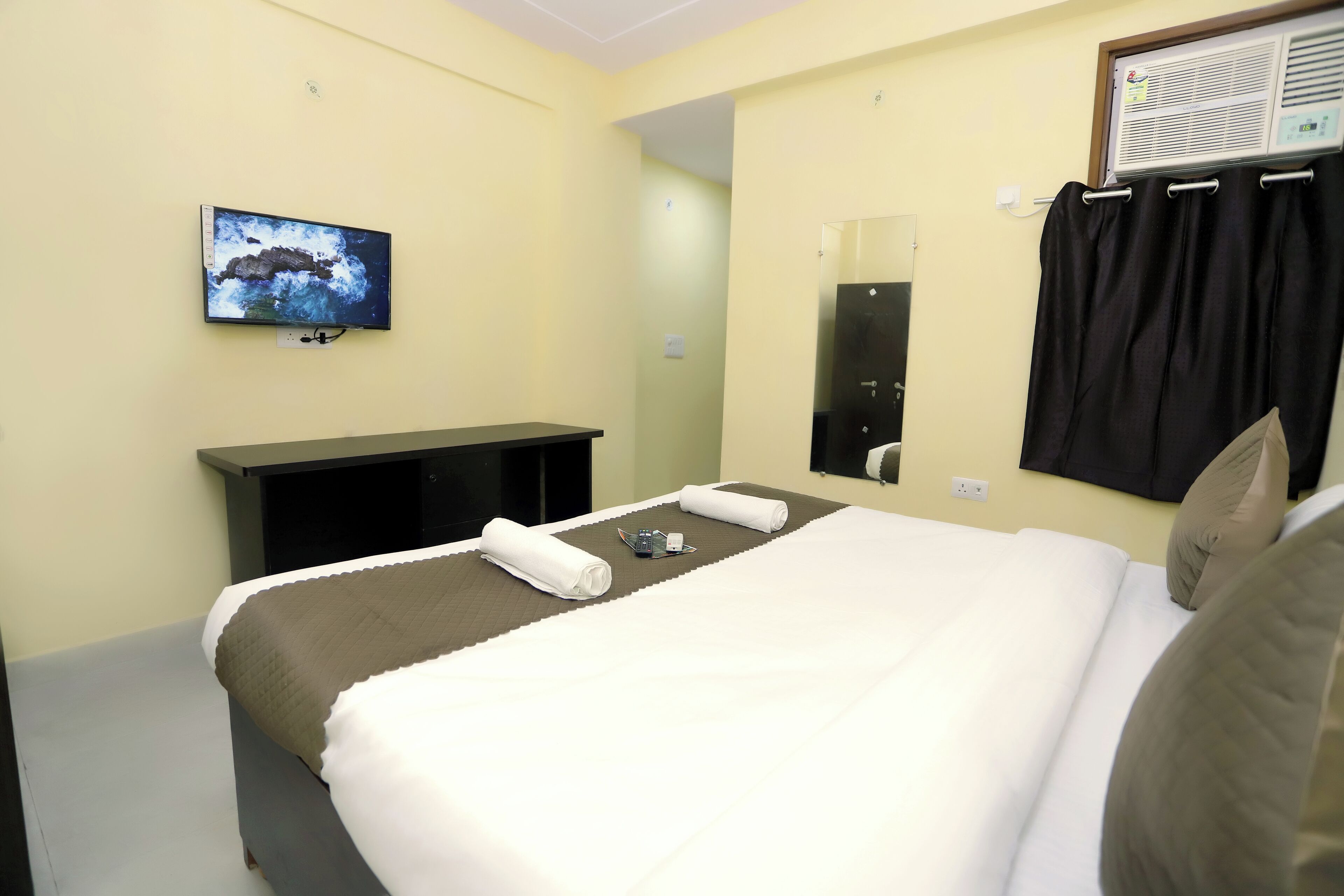 Een hotelkamer met een bed, een aan de muur bevestigde televisie, een nachtkastje, een spiegel en een raam met gordijnen.