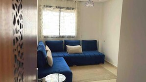 A living room with a blue sofa, a small round table, and a window with curtains. - Beautiful, spacious apartment (Témara)